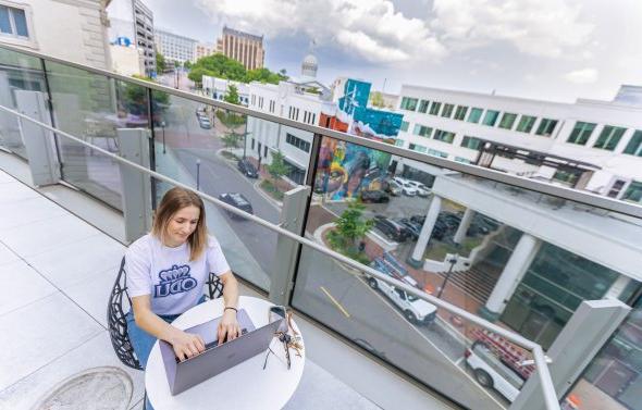 a female student with blond hair and a white odu t-shirt is sitting on a rooftop in a downtown area studying